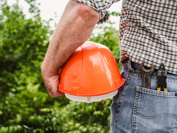 Attractive man in work clothes, holding construction helmet in his hands against the background of trees, blue sky and sunset. View from the back. Labor and employment concept - Foto, imagen