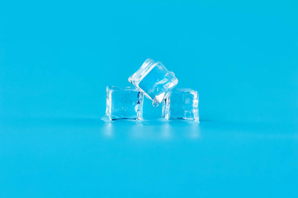 Melting ice on a blue background. A trickling drop from the central cube. Three ice cubes are lying in meltwater. - 写真・画像