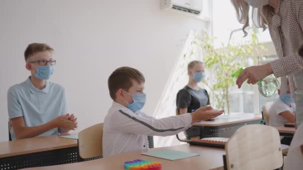 Tanár úr, gyerekek maszkkal az iskolában zárás után, fertőtlenítő kezek. iskolás fiú kinyújtja a kezét, miközben ül az asztalnál az osztályteremben, mint női tanár tölt néhány fertőtlenítő - Felvétel, videó