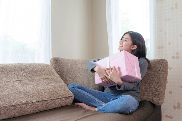 Aranyos tini női érzés boldogan és ölelés rózsaszín ajándék a kanapén, látszó előre kívül és mosolygós - Fotó, kép