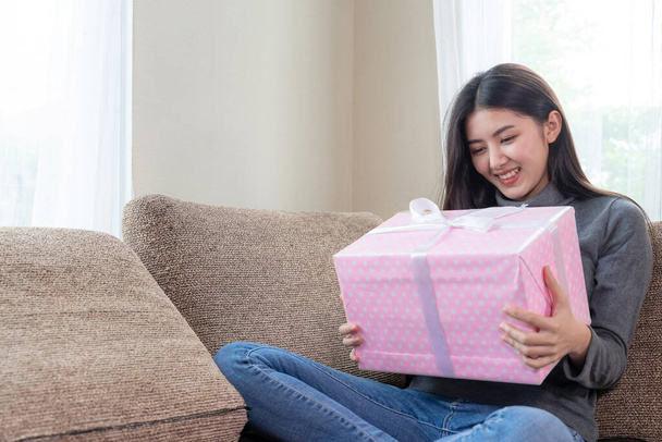 Söpö teini-ikäinen nainen tuntee onnellisesti ja halailee vaaleanpunaista lahjapakkausta sohvalla, etsii forwad ulkona ja hymyilee - Valokuva, kuva