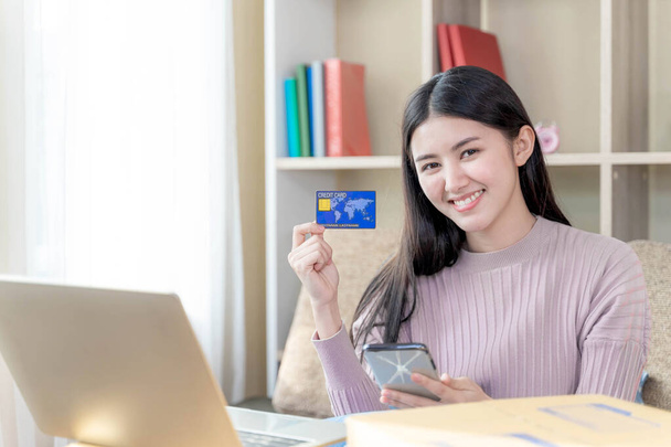 щаслива молода красива жінка сидить на дивані у вітальні вдома, вона посміхається і використовує кредитну картку для покупок онлайн - концепція онлайн-покупки та інтернет-банкінгу
  - Фото, зображення