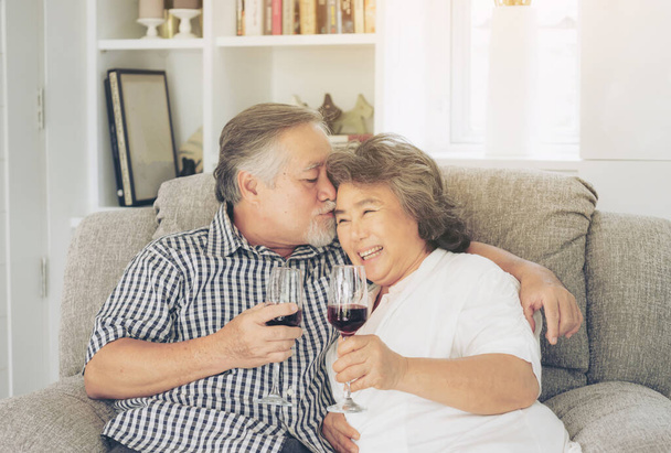 幸せな愛高齢者のカップルの笑顔は、ワイングラスを保持し、高齢者のカップルの男性と高齢者の女性はソファのリビングルームでリラックス結婚記念日を祝う-ライフスタイルのシニアコンセプト - 写真・画像