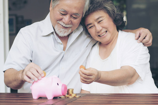 Fröhliches älteres Ehepaar spart Geld, indem es Münzen in Sparschwein und Münzstapel auf Holztisch legt, sie lächeln glücklich miteinander - Foto, Bild