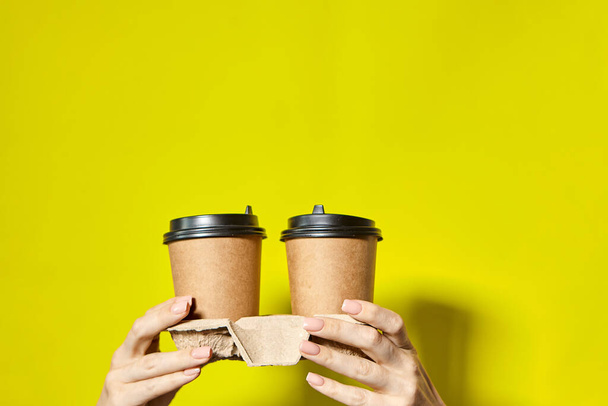 手は黒の蓋で2つのカップ茶色の紙を保持.2つのコーヒー特別オファーまたはプロモーション。黄色の画面の背景に2つのカップを保持手。紅茶やコーヒーを移動します。黒蓋の茶色い紙コップ. - 写真・画像