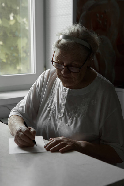 Starší žena píše doma na kus papíru. Koníčky důchodců. Vzdálená komunikace, posílání e-mailů. Proces psaní poznámky - Fotografie, Obrázek