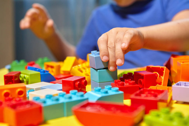 Ein schöner Junge spielt zu Hause mit Bauklötzen. Ein niedlicher lächelnder Junge spielt mit einem Lego-Konstrukteur mit vielen bunten Plastikklötzen im Raum und baut eine Stadt. Vorschulklassen. - Foto, Bild