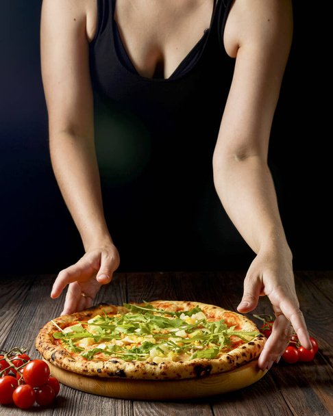 Tasty pizza on a black background with spices and vegetables. Womans hands reaching for pizza. Fresh arugula in hand. Italian cuisine concept. - Foto, imagen