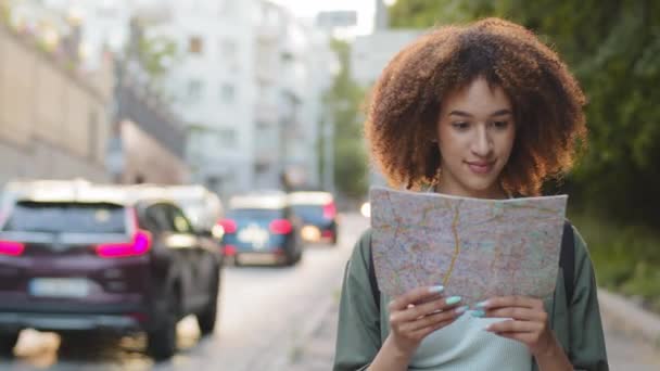 Χαρούμενη χαμογελαστή ελκυστική Αφροαμερικανή τουρίστρια με χάρτινο χάρτη στα χέρια την ηλιόλουστη μέρα του καλοκαιριού. Νεαρή μικτή φυλή θηλυκό ταξιδιωτικό blogger που ταξιδεύει στο κέντρο της πόλης χρησιμοποιεί οδηγό για τα αξιοθέατα - Πλάνα, βίντεο