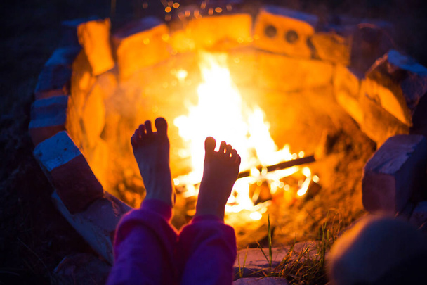 Bare feet of child by fire. Gatherings at night by campfire in open air in summer in nature. Family camping trip, gatherings around the campfire. Warm your feet, cold night - Photo, Image