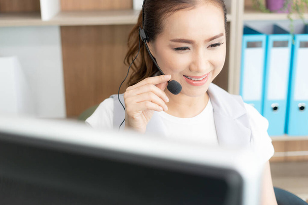 Hermosa chica asiática de atención al cliente con auriculares sonriendo durante una conversación telefónica - Foto, Imagen