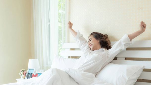 Hermosa mujer asiática sonriendo cara estirándose después de despertar disfrutando de la mañana soleada en la cama  - Foto, Imagen