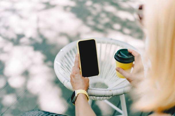 Cropped of womans hands holding yellow mobile phone with blank screen of gadget. Digital cellphone with mock up. Woman sitting cafe restaurant and drinking coffee. Copy space and mockup scene - Photo, Image