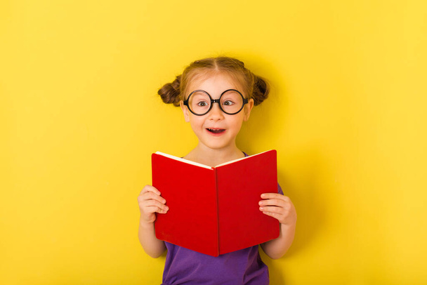 baby girl is holding book textbook and surprised. Little cute caucasian funny happy smile kid child on yellow background. children studio portrait. - 写真・画像