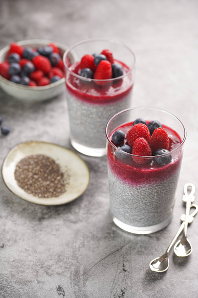 Two glasses with chia seeds pudding, raspberry jam and fresh raspberries and blueberries on a grey surface - Φωτογραφία, εικόνα