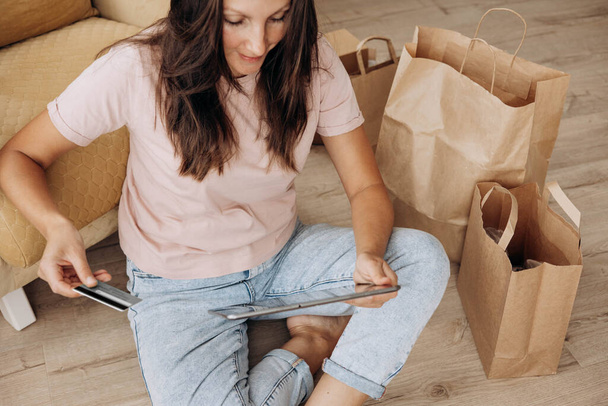 Femme assise sur le sol, tenant une carte bancaire de crédit et utilisant une tablette pour faire du shopping en ligne à la maison. Concentration sélective - Photo, image
