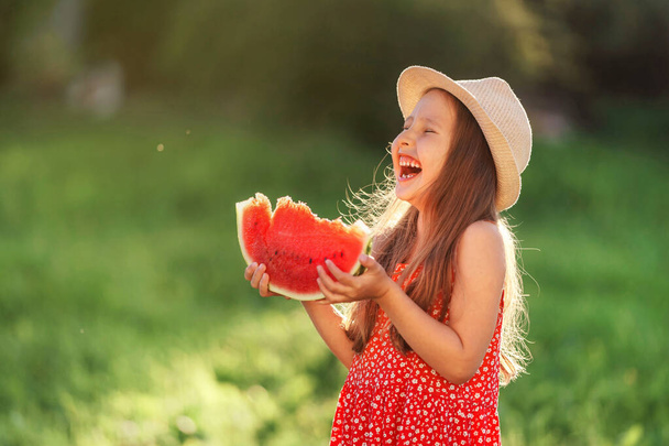 charming little girl in dress and hat, with light loose hair, is eating watermelon during summer holidays outdoors. cheerful happy child smiles and tries healthy fruit snack on sunny day, at sunset. - Φωτογραφία, εικόνα
