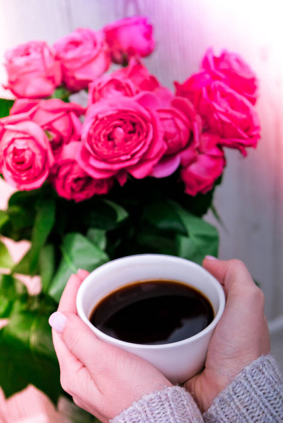 Kopje koffie en roze rozen. Vakantie concept, boeket rozen voor een verjaardag, boeket bloemen in de ochtend ontbijt. Vrouwelijke handen die cappuccino vasthouden. Valentijnsdag - Foto, afbeelding