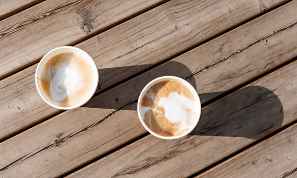 zwei Cappuccino in beigen Pappbechern auf holzgestreiftem Tisch draußen, Kaffee trinken, Freunde treffen, Licht und Schatten von oben - Foto, Bild