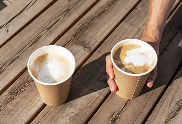 Twee cappuccino 's in beige papieren bekers op een houten gestreepte tafel buiten en een hand die een van hen vasthoudt, koffie drinkt, vrienden ontmoet - Foto, afbeelding