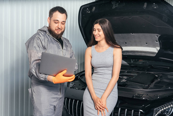 自動車修理工場でラップトップを使用して車両を検討しながら、若い顧客と通信する車の整備士 - 写真・画像