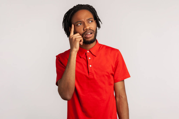 Portrait of young attractive bearded man with dreadlocks wearing red casual style T-shirt, thinking, has new idea, looking way with pensive expression. Indoor studio shot isolated on gray background. - Fotoğraf, Görsel