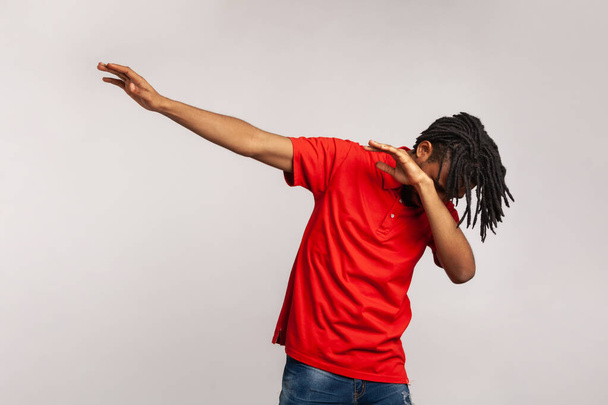 Portrait of young adult man with dreadlocks wearing red casual style T-shirt, standing in dab dance pose, internet meme, celebrating success. Indoor studio shot isolated on gray background. - Photo, image