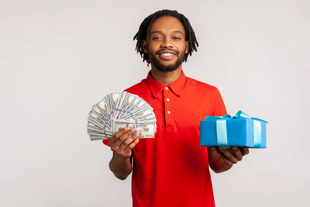 Happy man with dreadlocks and beard wearing red casual style T-shirt, holding present box, dollars banknotes, smiling to camera, cashback and bank loan. Indoor studio shot isolated on gray background. - Fotoğraf, Görsel