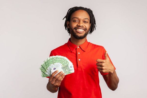 Positive man with dreadlocks wearing red casual T-shirt, holding bunch of euro banknotes and showing thumbs up gesture, sign of success, approval. Indoor studio shot isolated on gray background. - Photo, Image