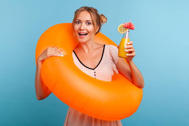 Excited woman with funny hair buns relaxing at summer resort, holding orange rubber ring and enjoying fresh fruit cocktail, laughing happily at vacation. Indoor studio shot isolated on blue background - Photo, Image