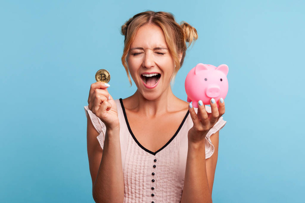 Excited pretty woman with blonde hair and wearing summer dress, yelling happily, holding piggy bank and gold crypto coin, growth bitcoin. Indoor studio shot isolated on blue background. - Photo, Image