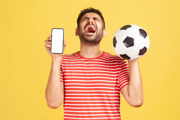 Extremely happy excited man with beard in striped t-shirt sincerely laughing holding white display smartphone and soccer ball, celebrating victory. Indoor studio shot isolated on yellow background - Photo, image