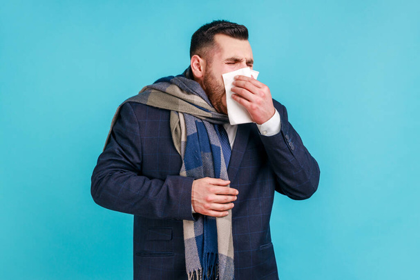 Cold and flu. Portrait of sick bearded young businessman in elegant suit standing, holding tissue on mouth and nose, being wrapped in warm scarf. Indoor studio shot isolated on blue background. - Φωτογραφία, εικόνα