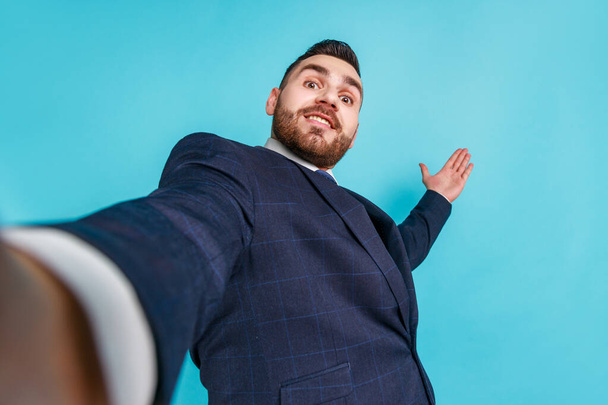 Portrait of young adult handsome businessman in elegant suit taking selfie, looking at camera POV and wave hand, point of view of photo. Indoor studio shot isolated on blue background. - Foto, afbeelding