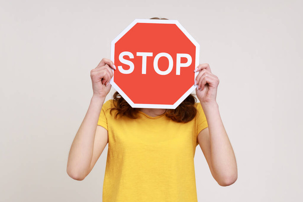 Way prohibited! Portrait of anonymous girl in yellow casual T-shirt covering face with stop symbol, holding red traffic sign, forbidden access. Indoor studio shot isolated on gray background. - 写真・画像