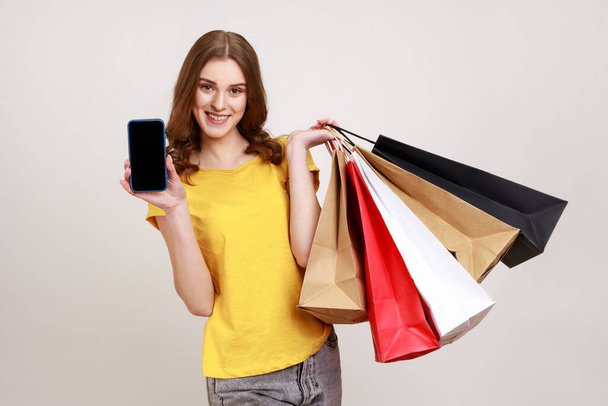 Portrait of happy smiling teenager girl in yellow T-shirt holding cell phone and shopping bags, advertising online store on mobile device. Indoor studio shot isolated on gray background. - Photo, Image