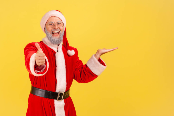 Glad happy elderly man with gray beard wearing santa claus costume presenting copy space for advertisement on his palm, showing thumb up, recommend. Indoor studio shot isolated on yellow background. - Zdjęcie, obraz