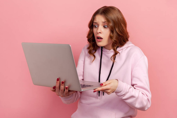 Portrait of curly haired young female in hoodie looking at laptop display with big eyes, shocked with job requirements, deadline. Indoor studio shot isolated on pink background - Foto, immagini
