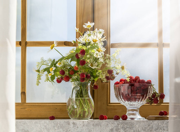 Au nez avec des camomilles et des branches de framboises et framboises dans un vase sur le rebord de la fenêtre. - Photo, image