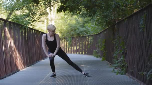 Középkorú szőke fehér nő sportoló hölgy visel fekete melegítő áll a hídon a városi parkban kanyarokban térd guggol nyújtózik izmok lábak csinál nyújtás edzés sport szabadban rugalmasság - Felvétel, videó