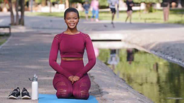 Boldog sportos fekete nő visel sportruházat ül jóga szőnyeg a szabadban nézi a kamerát, vár edzés után, vagy pihentető edzés után. Plusz méret gyönyörű afro-amerikai modell nyári parkban - Felvétel, videó