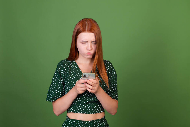 Yeşil arka planda genç kızıl saçlı kadın cep telefonuyla mesajlaşan sinirli sinirli, ciddi hayal kırıklığına uğramış kadın. - Fotoğraf, Görsel
