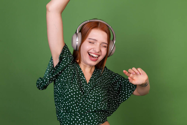 Joven mujer de pelo rojo sobre fondo verde con auriculares inalámbricos feliz moviéndose bailando - Foto, Imagen