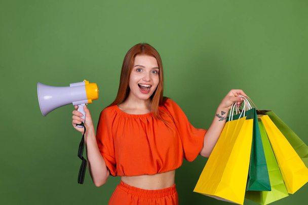 Jovem mulher de cabelo vermelho na blusa laranja casual no fundo verde com sacos de compras gritando em megafone - Foto, Imagem