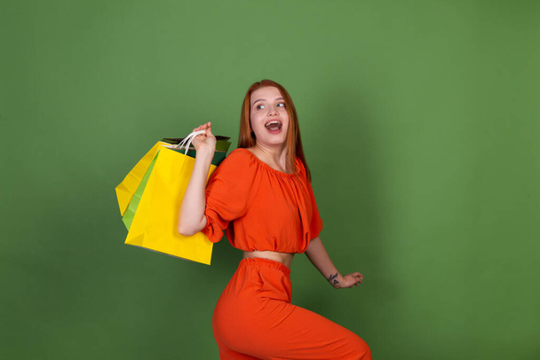 Jovem mulher de cabelo vermelho na blusa laranja casual no fundo verde com sacos de compras feliz positivo animado alegre - Foto, Imagem