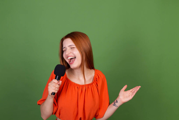 Mladý ryšavý vlasy žena v ležérní oranžová halenka na zeleném pozadí drží mikrofon zpěv  - Fotografie, Obrázek