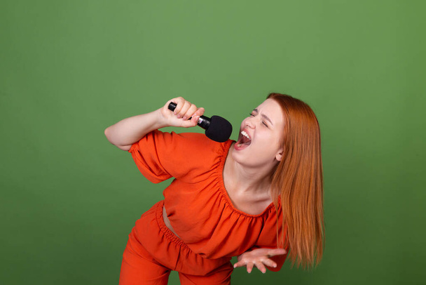 Mujer de pelo rojo joven en blusa naranja informal sobre fondo verde sosteniendo el micrófono cantando  - Foto, Imagen
