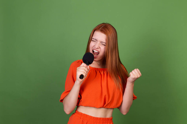 Νεαρή κόκκινα μαλλιά γυναίκα σε casual πορτοκαλί μπλούζα σε πράσινο φόντο κρατώντας μικρόφωνο τραγουδώντας  - Φωτογραφία, εικόνα