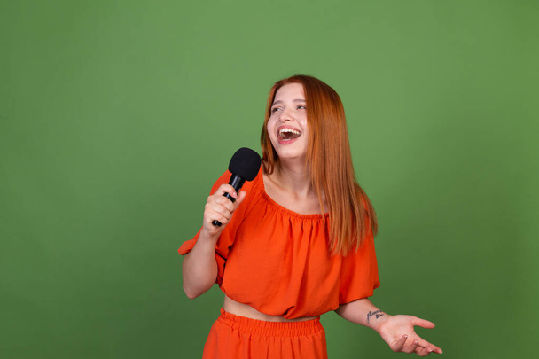 Νεαρή κόκκινα μαλλιά γυναίκα σε casual πορτοκαλί μπλούζα σε πράσινο φόντο κρατώντας μικρόφωνο τραγουδώντας  - Φωτογραφία, εικόνα