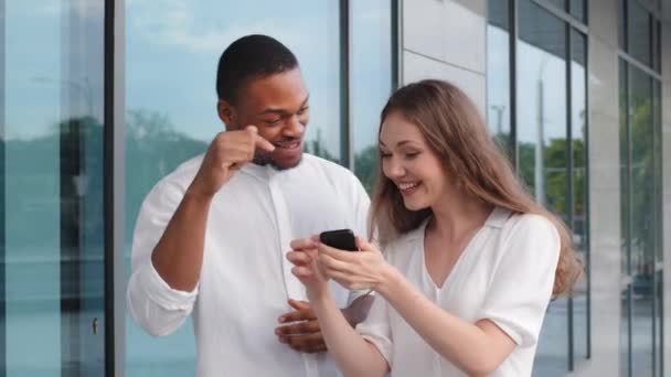 Muotokuva monirotuinen pari afro amerikkalainen mies afrikkalainen mies ja valkoihoinen nainen tyttö kaksi ystävää seistä ulkona katsomassa näytön puhelimen video verkossa nauraa hymyillen mobiilisovelluksella älypuhelimeen - Materiaali, video